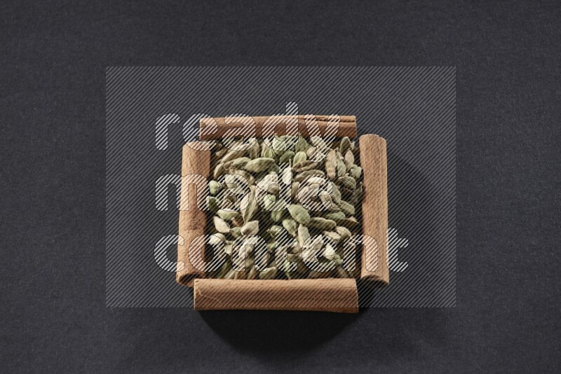 أربعة من أعواد القرفة مكونة شكل مربع بداخلها حبوب الهيل علي خلفية سوداء