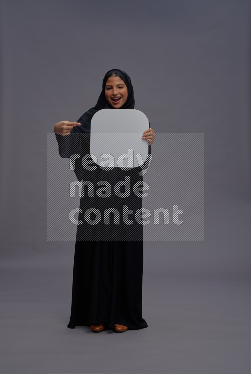 Saudi woman wearing Abaya standing holding social media sign on gray background