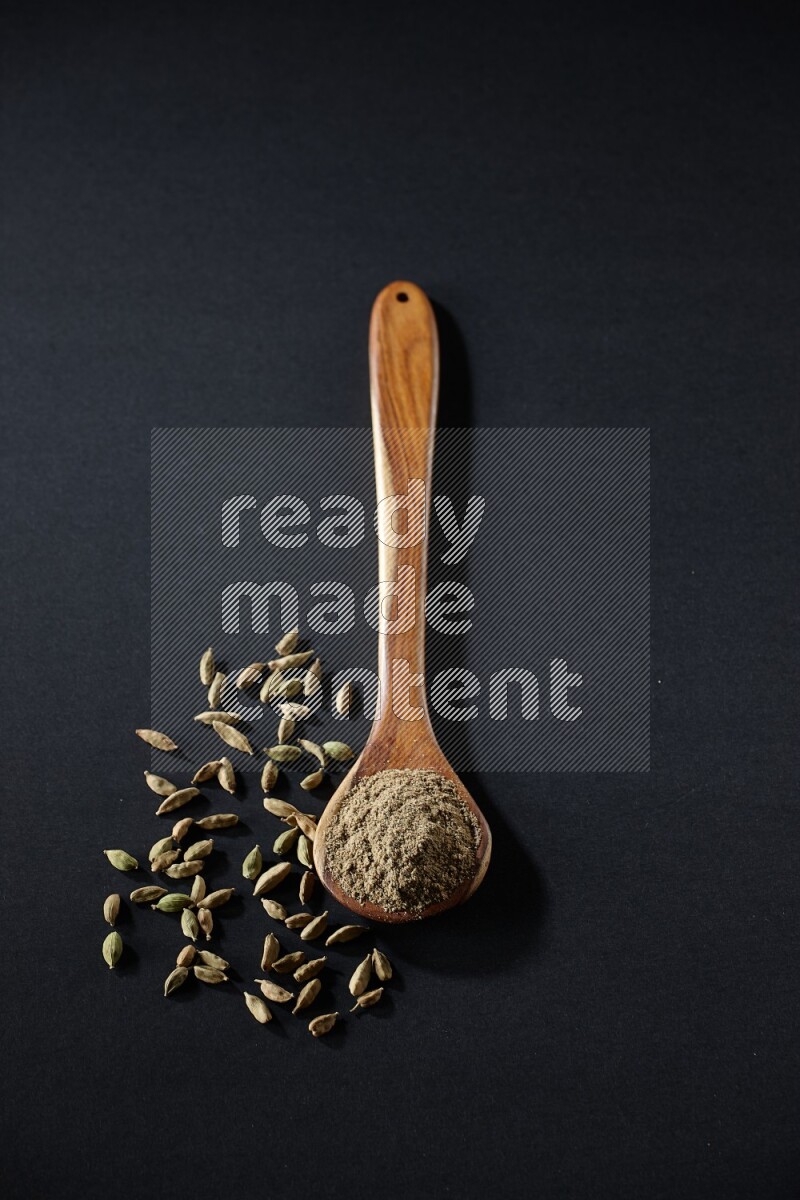 A wooden ladle full of cardamom powder and cardamom seeds beside it on black flooring