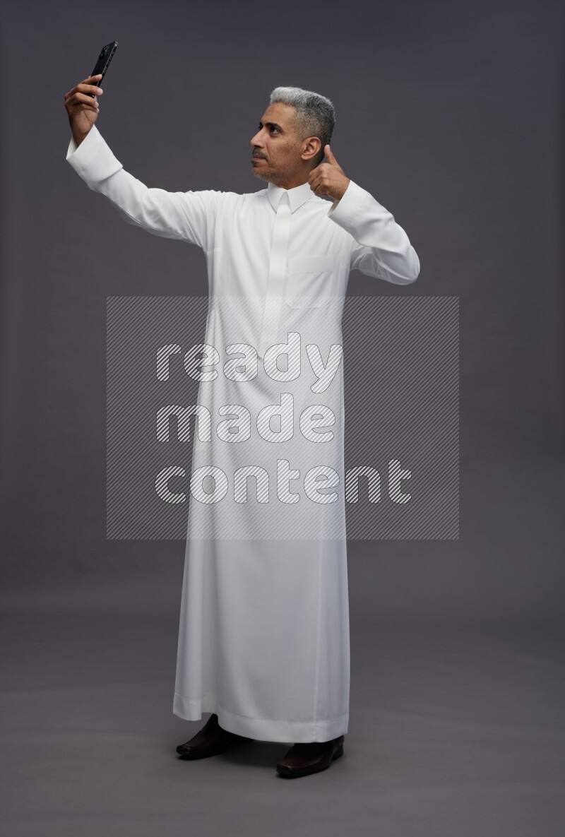 Saudi man wearing thob standing taking selfie on gray background