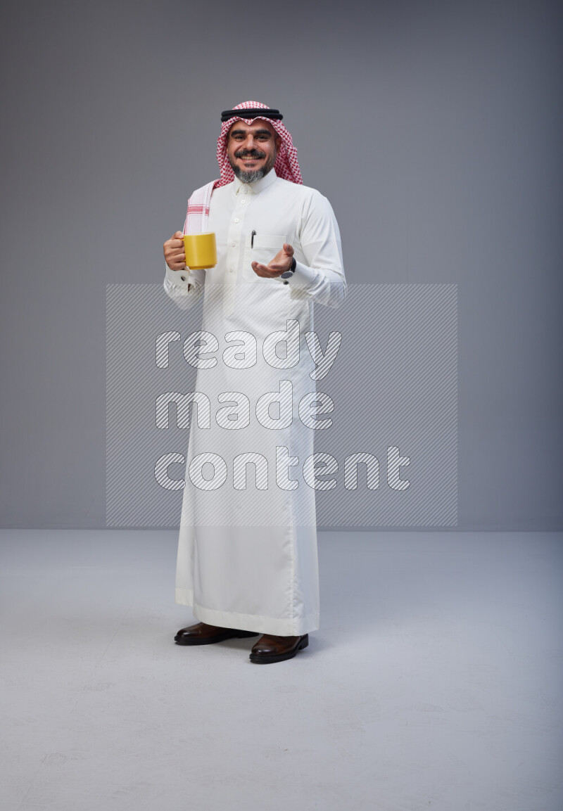 Saudi man Wearing Thob and red Shomag standing holding mug on Gray background