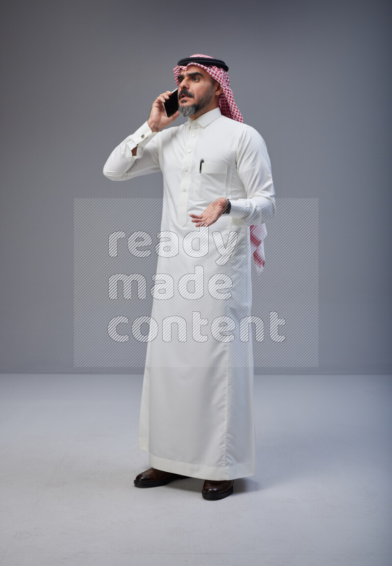 Saudi man Wearing Thob and red Shomag standing talking on phone on Gray background