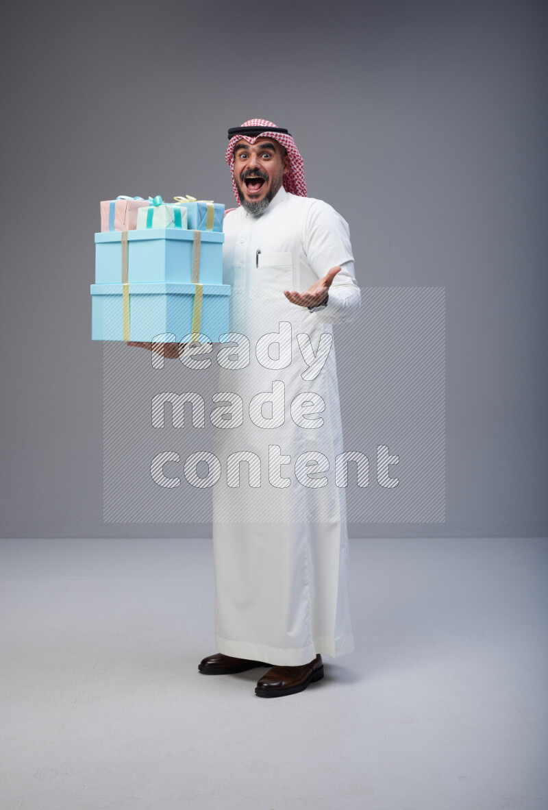 Saudi man Wearing Thob and red Shomag standing holding gift box on Gray background