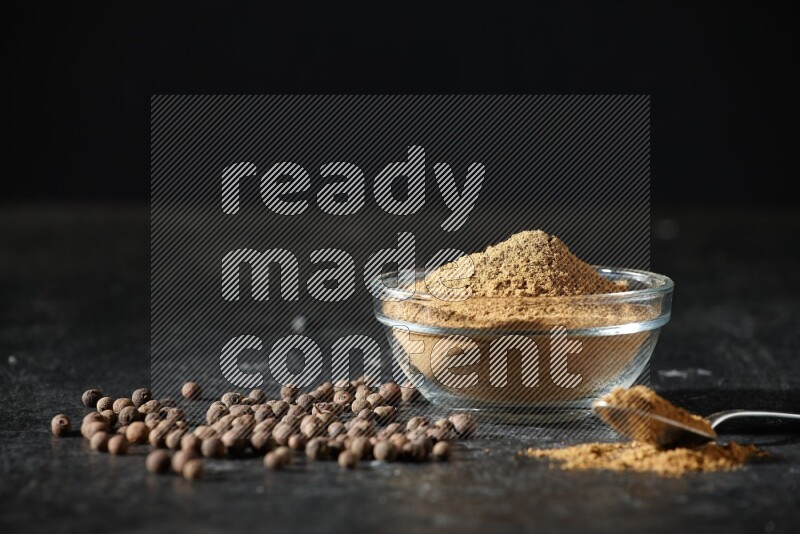 A glass bowl and metal spoon full of allspice powder and whole balls spreaded on a black flooring