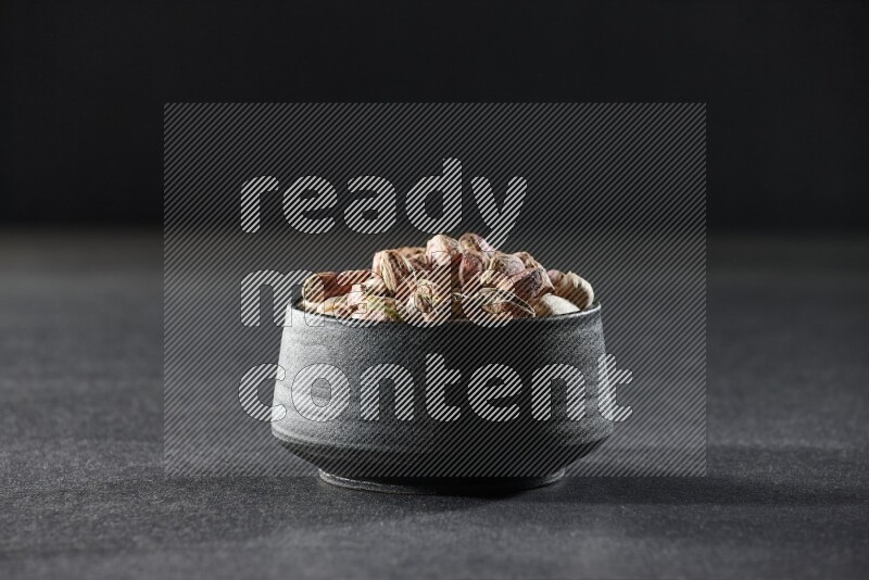 A black pottery bowl full of peeled pistachios on a black background in different angles