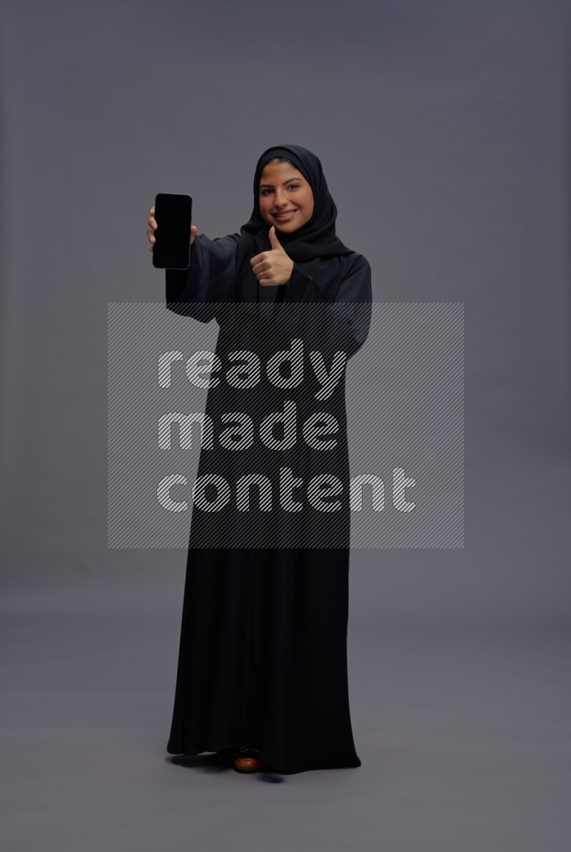 Saudi woman wearing Abaya standing showing phone to camera on gray background