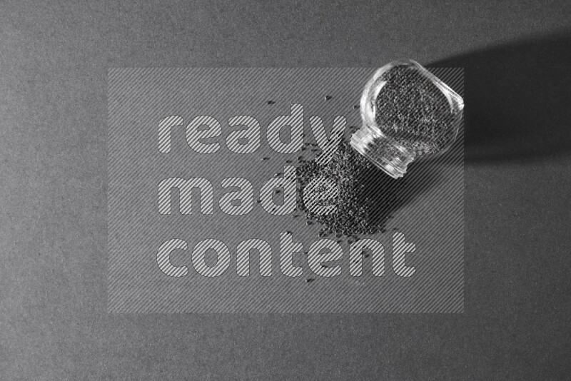 A flipped glass spice jar full of black seeds and the seeds spread out on a black flooring