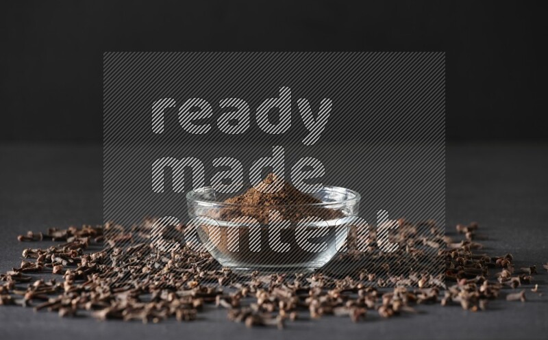 وعاء زجاجي ممتلئ ببودرة القرنفل مع حبوب القرنفل المتناثرة على أرضية سوداء