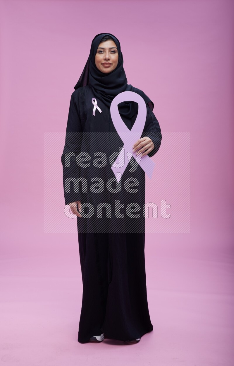 Saudi woman wearing pink ribbon on abaya standing holding awareness ribbon on plain pink background