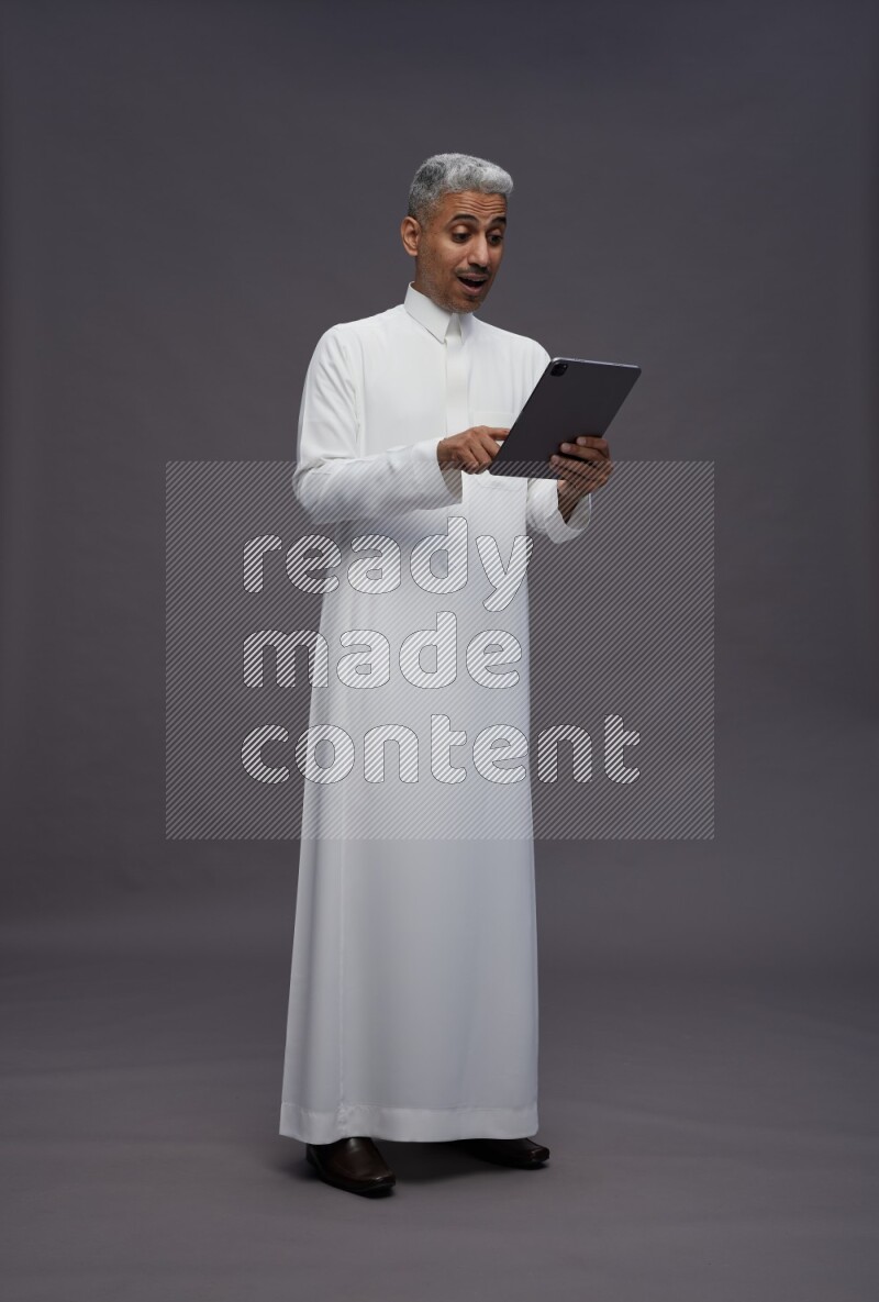 Saudi man wearing thob standing working on tablet on gray background