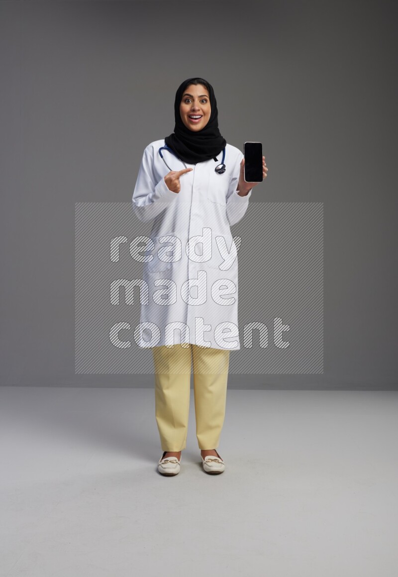 Saudi woman wearing lab coat with stethoscope standing showing phone to camera with sign in the back on Gray background