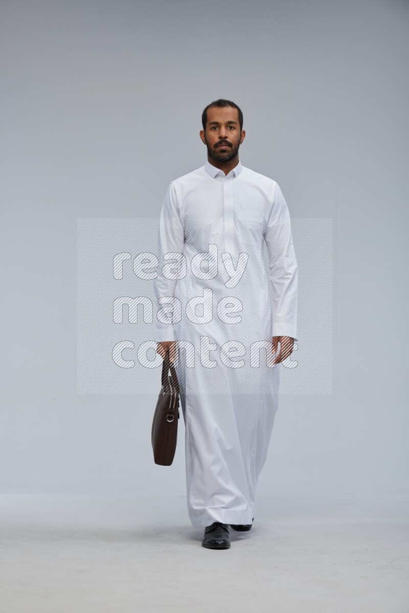Saudi man wearing thob standing holding suitcase on gray background