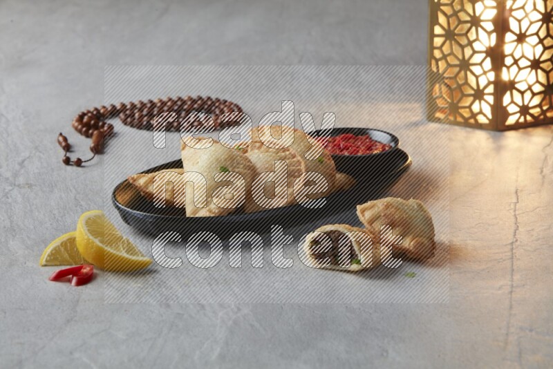 Five fried sambosas in an oval shaped black plate, beside a cut meat sambosa, a brown misbaha and a golden lantern on a gray background