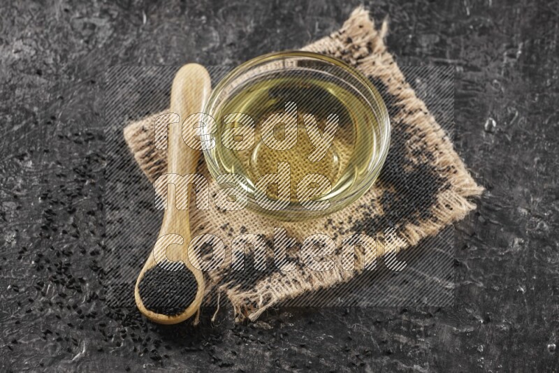 وعاء زجاجي ممتلئ بزيت حبة البركة مع ملعقة خشبية بها بذور حبة البركة مع بعض البذور المتناثرة علي قطعة من القماش علي خلفية سوداء