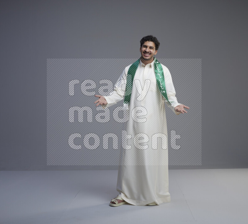 A Saudi man standing wearing thob and Saudi flag scarf on gray background
