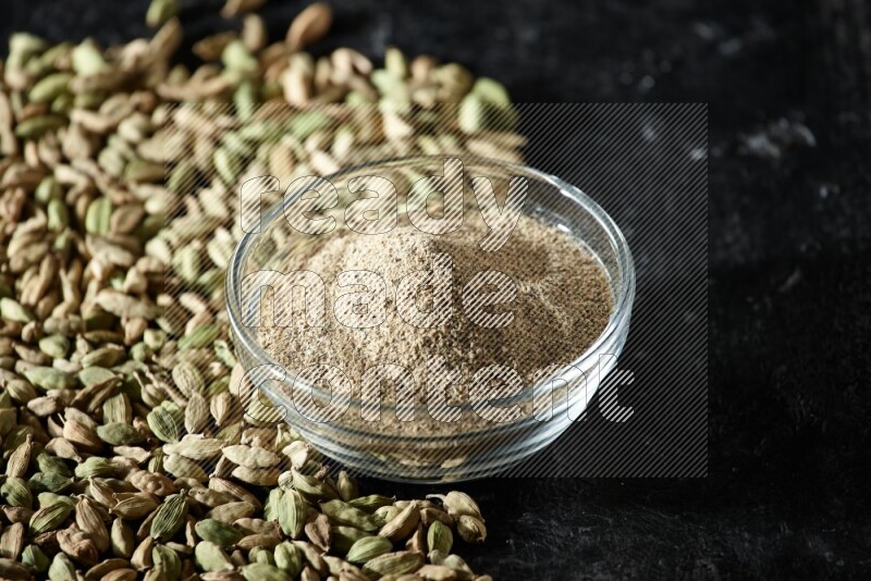 A glass bowl full of cardamom powder and cardamom seeds spreaded beneath the bowl on textured black flooring