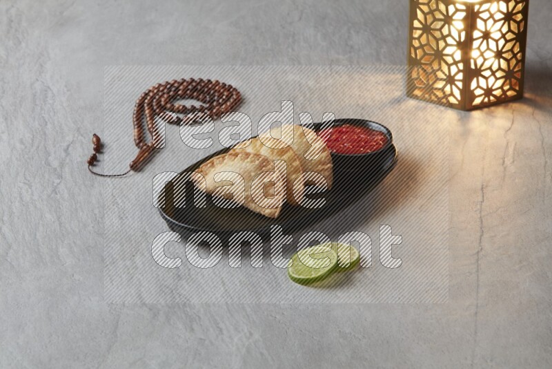 Three fried sambosas in an oval shaped black plate and a red sauce in a black round ramekin with a brown misbaha and a golden lantern on the side on a gray background