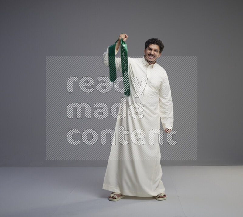 A Saudi man standing wearing thob holding Saudi flag scarf on gray background