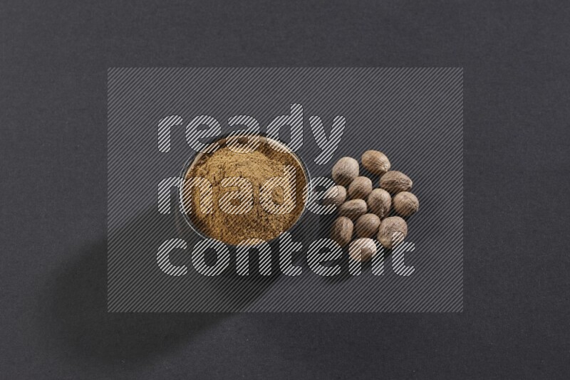 A black pottery bowl full of nutmeg powder with whole seeds beside it on a black flooring