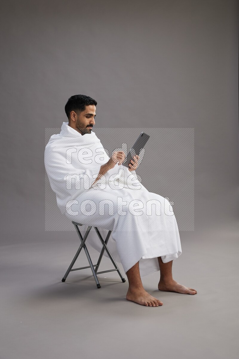 A man wearing Ehram sitting on chair holding a tablet on gray background