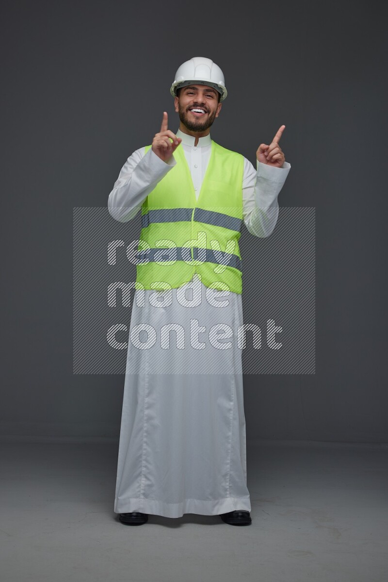 A Saudi man wearing Thobe with a yellow safety vest and white helmet standing and pointing different angles eye level on a gray background