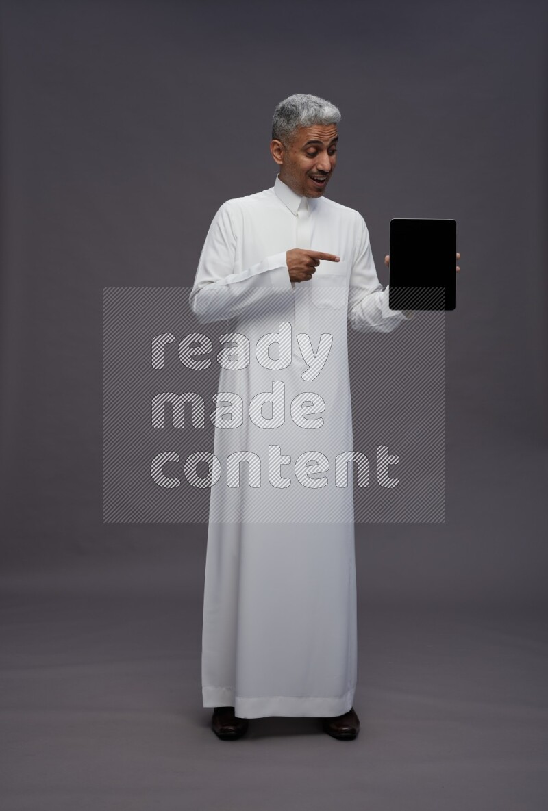 Saudi man wearing thob standing showing tablet to camera on gray background