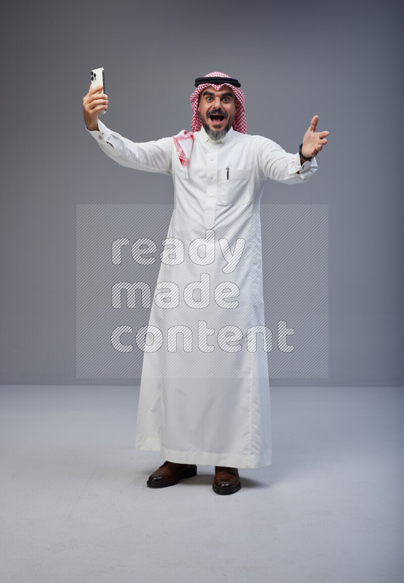 Saudi man Wearing Thob and red Shomag standing taking selfie on Gray background