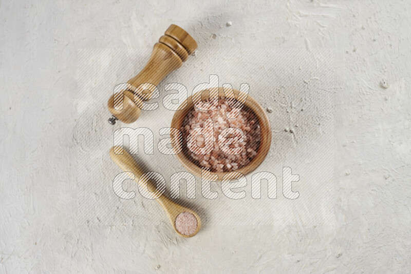A wooden bowl and spoon filled with coarse pink himalayan salt and a wooden grinder beside them on white background
