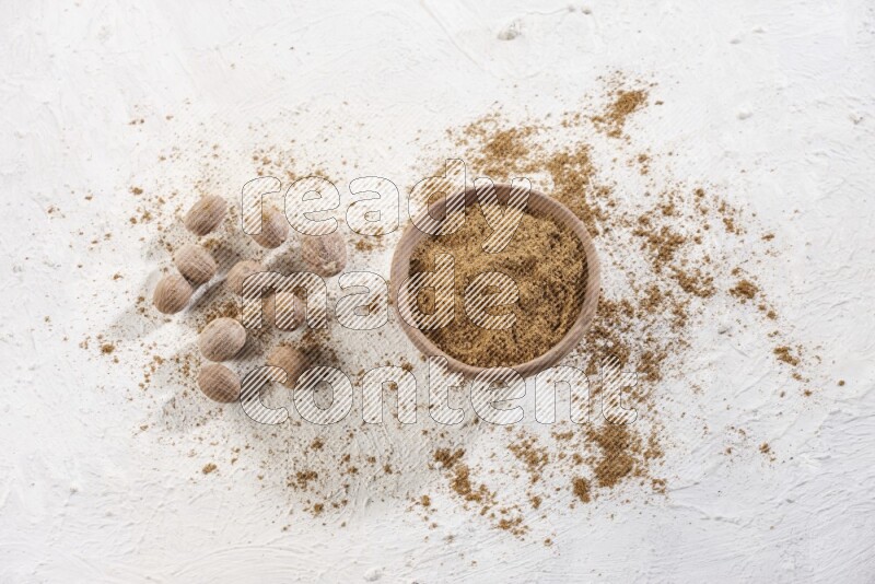 A wooden bowl full of nutmeg powder with whole seeds and sprinkled powder beside it on a textured white flooring