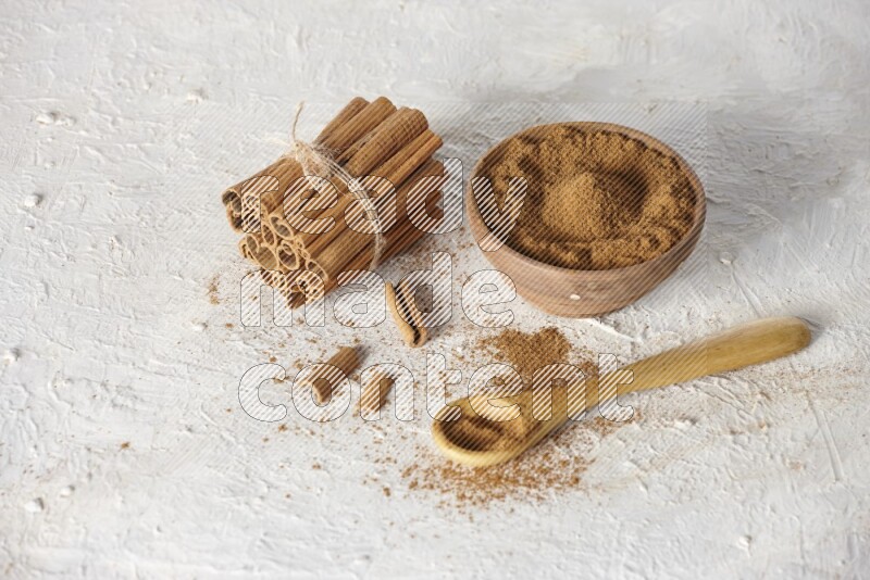 Cinnamon sticks stacked and bounded beside a wooden bowl full of cinnamon powder and a wooden spoon full of powder on white background