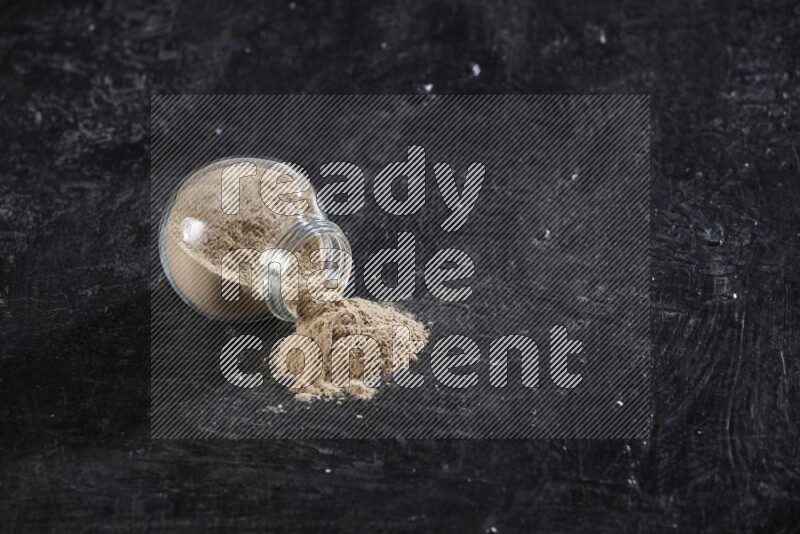 A glass spice jar full of garlic powder flipped over with the powder came out on a textured black flooring