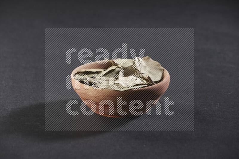 A wooden bowl filled with dried bay leaves on black flooring