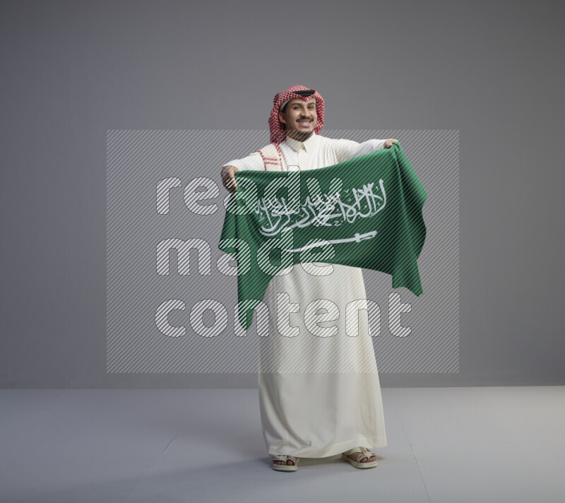 A saudi man standing wearing thob and red shomag holding big saudi flag on gray background