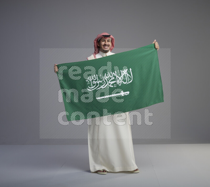 A saudi man standing wearing thob and red shomag holding big saudi flag on gray background