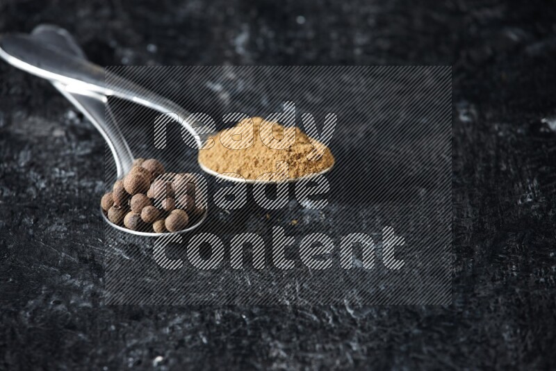 ملعقتان معدنيتان ممتلئتان ببودرة بهار الفلفل الحلو وحبوبه على خلفية سوداء