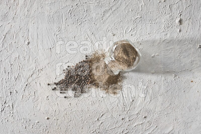 Flipped glass spice jar full of black pepper powder and paper beads beside it on a textured white flooring