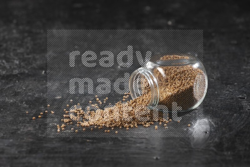 A glass spice jar full of mustard seeds and jar is flipped and seeds spread out on a textured black flooring