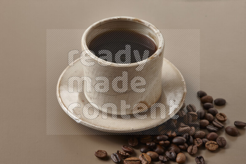 A beige pottery cup of coffee surrounded by roasted coffee beans on beige background