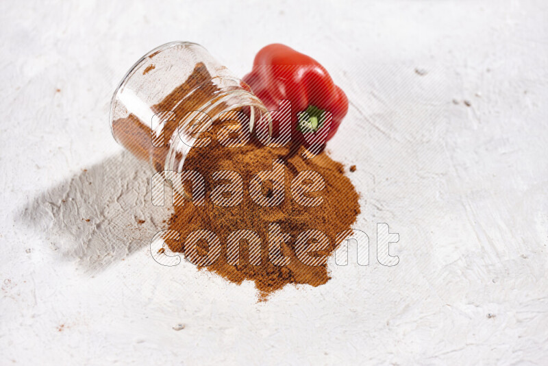 A glass jar full of ground paprika powder flipped with some spilling powder on white background