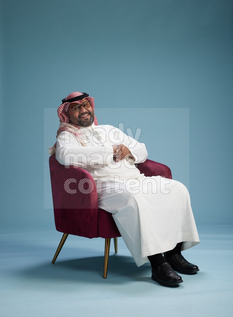 Saudi Man with shimag sitting on chair Interacting with the camera on blue background