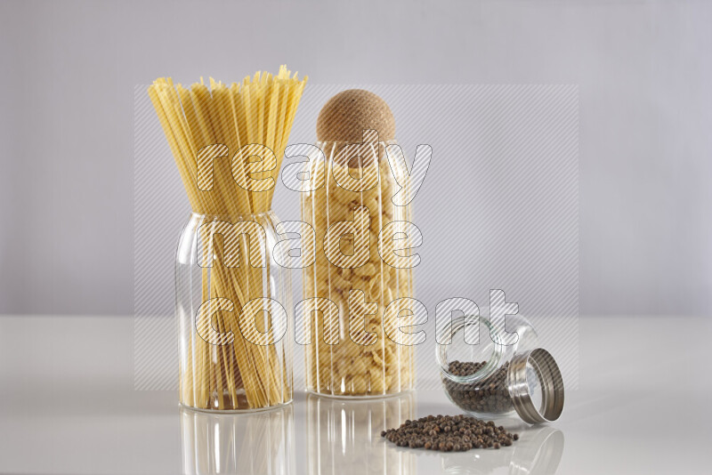 Raw pasta in glass jars with black peppers on light grey background