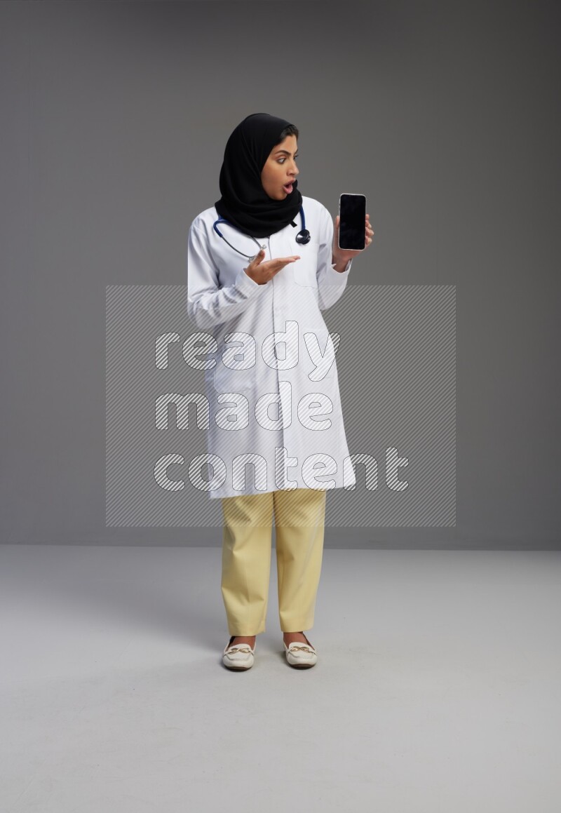 Saudi woman wearing lab coat with stethoscope standing showing phone to camera with sign in the back on Gray background