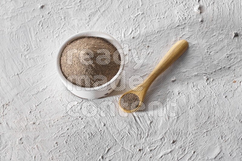 A pottery white bowl full of black pepper powder and wooden spoon full of powder on textured white flooring
