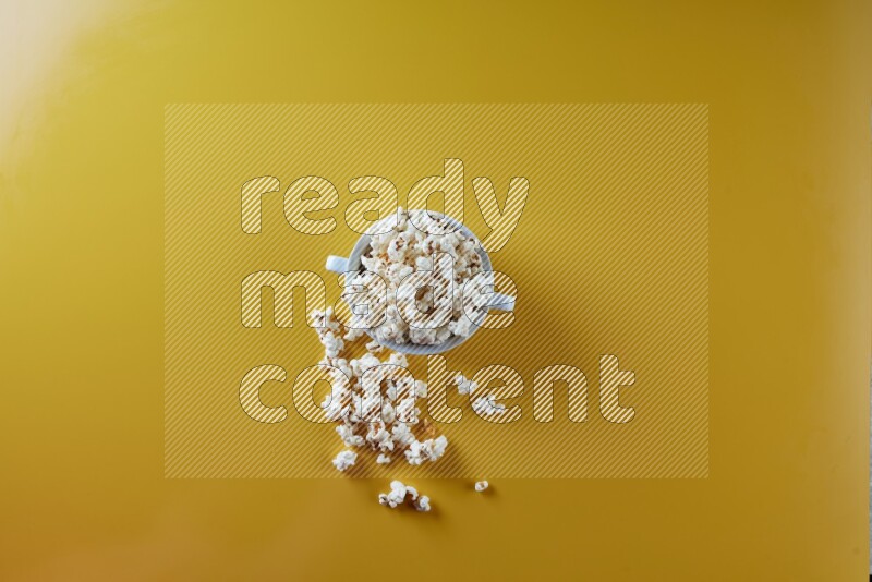 A white ceramic bowl full of popcorn with popcorn beside it on a yellow background in a top view shot