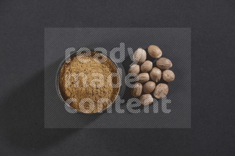 A black pottery bowl full of nutmeg powder with whole seeds beside it on a black flooring