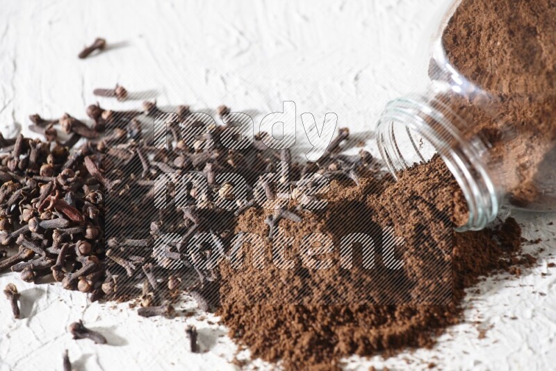 وعاء زجاجي للتوابل مقلوب ممتلئ ببودرة القرنفل وتناثرت البودرة منه مع حبوب القرنفل المنتشرة على أرضية بيضاء