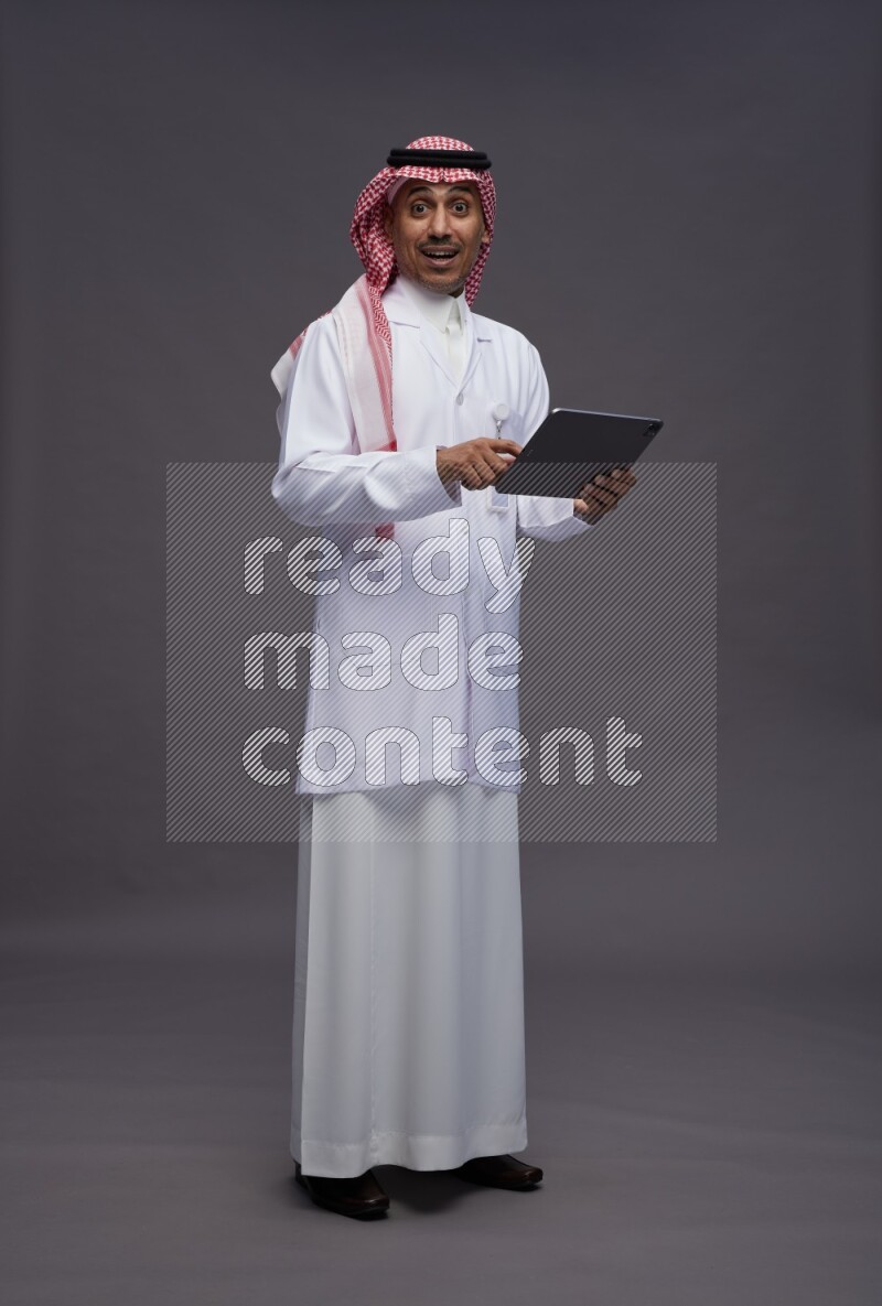 Saudi man wearing thob with lab coat and shomag with pocket employee badge standing working on tablet on gray background