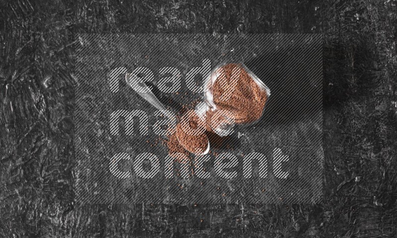 وعاء زجاجي مقلوب ممتلئ ببذور حب الرشاد مع بعض الحبوب المتناثرة منه مع ملعقة معدنية ممتلئة بالبذور على أرضية سوداء