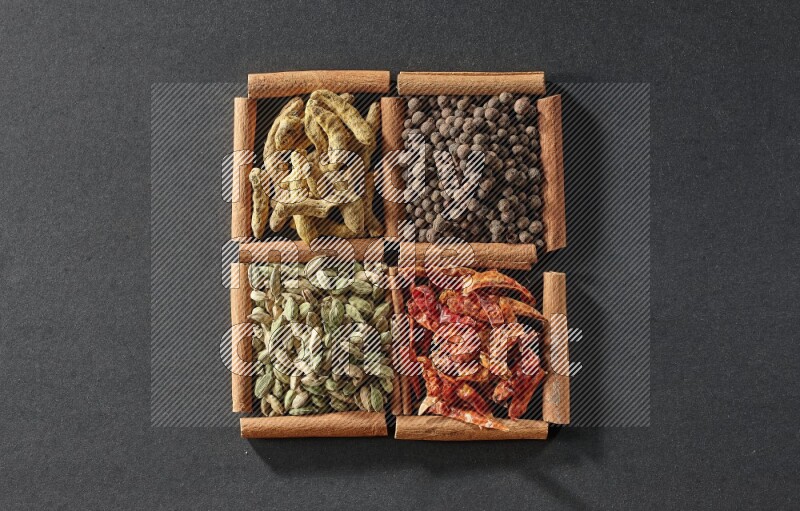 4 مربعات من عيدان القرفة ممتلئة بحبوب الهيل والفلفل الحار وبهار الفلفل الحلو وأصابع الكركم على أرضية سوداء