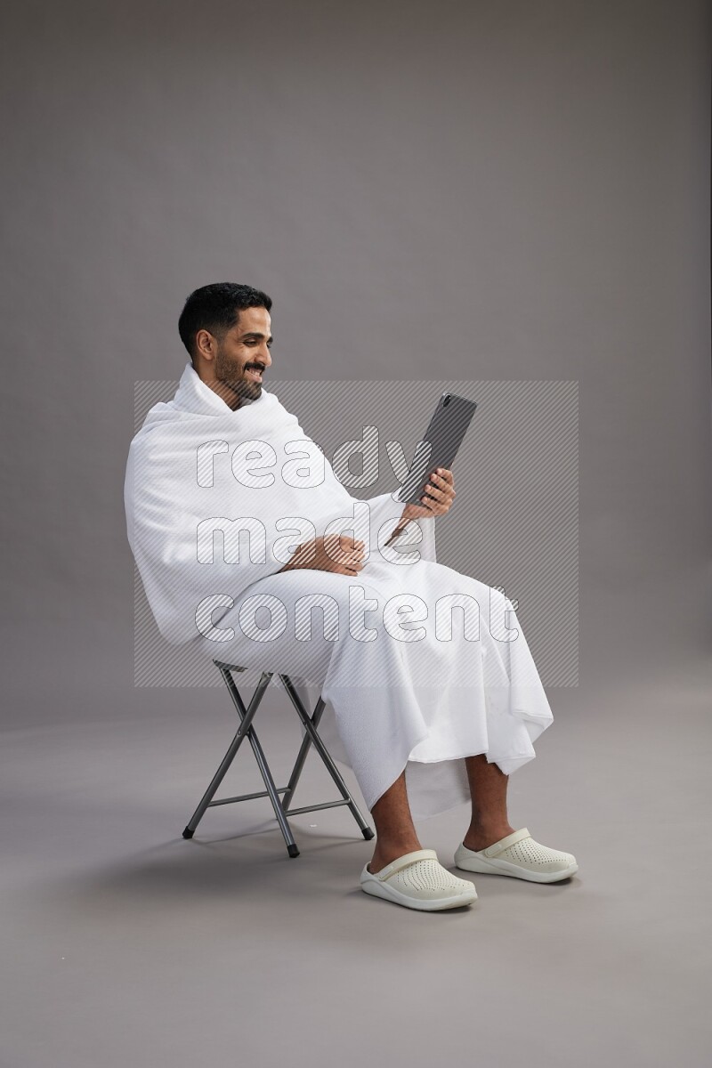 A man wearing Ehram sitting on chair holding a tablet on gray background
